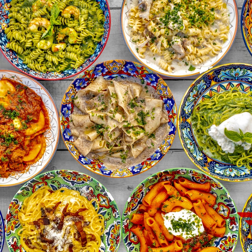 An overhead view of eight colorful bowls filled with various Italian pasta dishes, including pesto, carbonara, and ravioli.