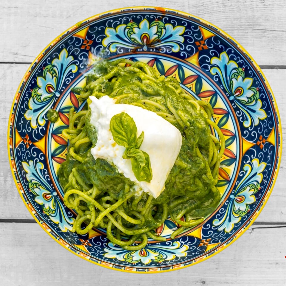 Green pesto pasta with a dollop of burrata cheese and a basil leaf on a colorful plate.